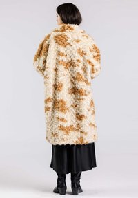 Konstpälsjacka med ett krämvitt och brunt mönster, oversized passform, texturerad yta och hög krage. Bärs över en svart kjol och stövlar.