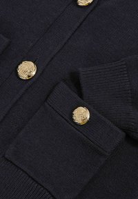 Marinblå cardigan i strukturerad stickad tyg, med dekorativa knappar i guld med ett emblem, samt ribbade muddar och fickkanter.