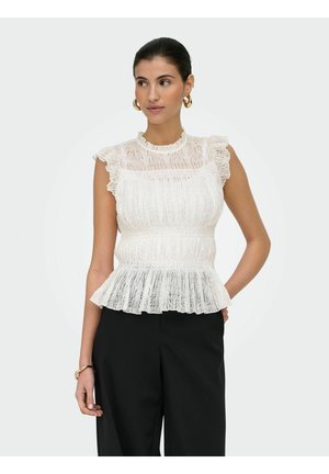 Kobieta nosząca białą koronkową bluzkę bez rękawów z falbankami oraz czarne szerokie spodnie, uzupełniona złotymi kolczykami kołami i bransoletką.