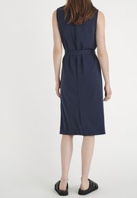 Granatowa sukienka bez rękawów z paskiem w talii, o długości do kolan i rozcięciem z tyłu dla swobody ruchów. Gładka faktura materiału.