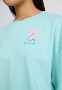 Tricou din bumbac light blue, cu mâneci scurte, având un design floral roz și logo-ul Columbia pe pieptul stâng. Textură moale.