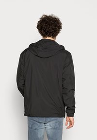 Svart hettegenser med en glatt, blank tekstur, elastiske mansjetter og kanten. Båret over lyseblå jeans, sett fra baksiden.