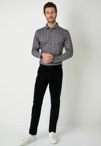 Heren overhemd met een patroon in donkere tinten en een bloemenmotief, gecombineerd met zwarte corduroypantalons en witte sneakers, staande tegen een effen achtergrond.