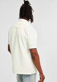 Korte mouw shirt in lichtcrème, gestructureerde stof. Heeft een kraag en een afgeronde zoom, met een knoopdetail op de achterkant.