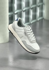 Hvite atletiske joggesko med strukturert finish, grå aksenter og en blå hælflik. Sålen har et mønstret mønster for grep.