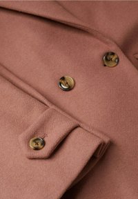 Gros plan sur un manteau en laine rose poussiéreux avec des boutons en écaille et un détail de poignet plié.
