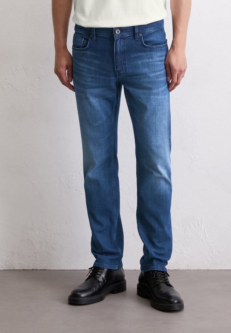Jeans en denim bleu avec une coupe droite, dotés d'un design à cinq poches, fermeture à bouton et un léger délavage sur les jambes. Associés avec des chaussures noires.
