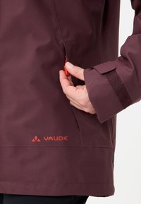 Giacca impermeabile color bordeaux con tasca laterale con zip e polsini in velcro. Presenta il marchio rosso "VAUDE" vicino all'orlo. Tessuto liscio e resistente.