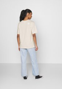 T-shirt in cotone beige con scollatura rotonda e maniche corte, mostrata da dietro, con un piccolo logo bianco nella parte superiore della schiena. I jeans in denim azzurro chiaro e le sneakers nere completano il look.