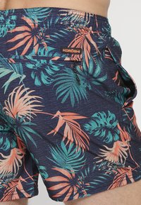 Costum de baie din țesătură albastră cu un model de frunze tropicale în nuanțe de teal și corai. Dispune de un buzunar cu fermoar și o etichetă cu logo pe talie.