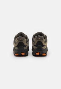 Baskets de sport vert olive et noires avec une tige texturée, des accents orange contrastants et une semelle rembourrée dotée de motifs de semelle distincts.