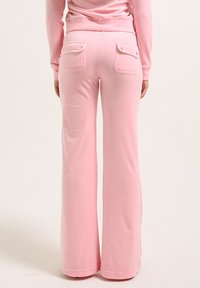 Des pantalons de survêtement roses à jambe évasée avec deux poches arrière à boutons-pression, une finition texturée douce et une taille élastique.