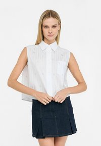Camicia bianca senza maniche con colletto, caratterizzata da linee verticali argento e una tasca sul petto, abbinata a una mini gonna di jeans scuri.