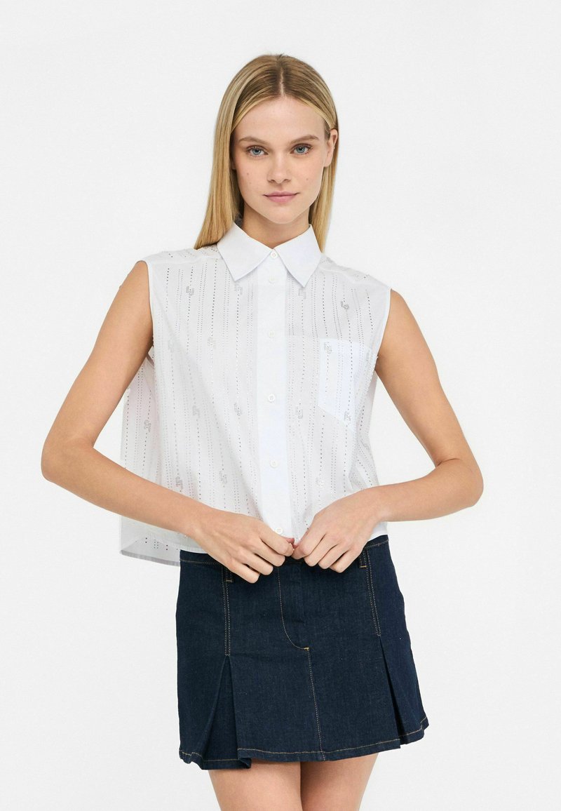 Camicia bianca senza maniche con colletto, caratterizzata da linee verticali argento e una tasca sul petto, abbinata a una mini gonna di jeans scuri.