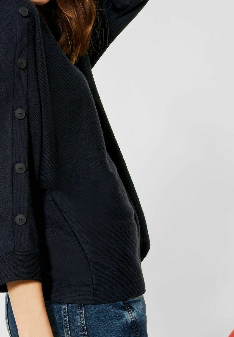 Mörkblå tröja med en texturerad yta, med stora svarta knappar på sidan och trekvartslånga ärmar.