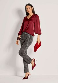 Bordeauxfarbene Satinbluse mit Puffärmeln, kombiniert mit grauen gemusterten Hose und kastanienbraunen High Heels. Die Accessoires umfassen einen Kettengürtel und eine rote Clutch.