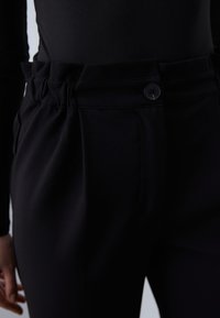 Pantalon noir en tissu lisse, avec une fermeture à bouton à la taille, un devant plissé et une ceinture élastique pour un confort accru.