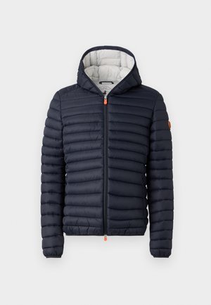 Mørkeblå quiltet hættejakke med frontlynlås, orange lynlåskroge og orange logo-patch på venstre ærme.