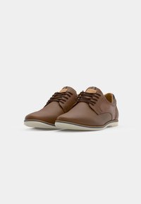Chaussures en cuir marron avec une texture lisse, bout arrondi et semelle blanche avec une fine bande bleue. Lacets et petits logos visibles.