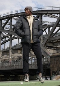 Chaqueta negra de plumas, forro de polar crema debajo, pantalones oscuros, gorro gris y zapatillas deportivas grises, con un fondo de puente industrial.