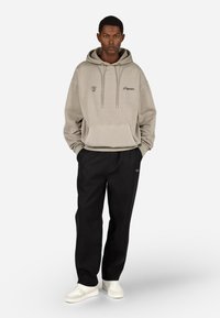 Gaišas pelēks kapucīna džemperis ar priekšējo kabatu un logotipu. Saskaņots ar melniem biksēm. Modelis valkā baltas sporta kurpes. Vienkāršs, ikdienišķs dizains.