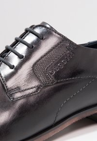 Chaussure en cuir noir à finition lisse, panneau latéral texturé, six œillets et logo de la marque embossé sur le cuir. Design minimaliste.