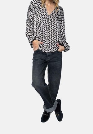 Femme portant un chemisier à pois noirs et blancs, un jean sombre retroussé, des mocassins noirs, debout les mains dans les poches et jambes croisées.