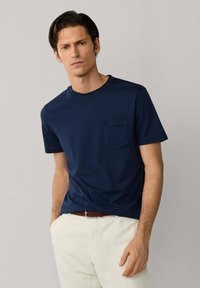 Hombre con camiseta azul marino de manga corta con bolsillo y pantalones blancos, de pie con las manos en los bolsillos contra un fondo claro y liso.