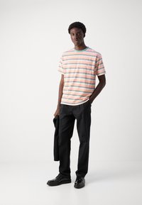 Stribet t-shirt i pink og turkis, med korte ærmer og en brystlomme, parret med sorte bukser og sko.