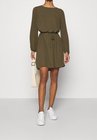 Olivgrön klänning med långa veckade ärmar, midja med resår och längd ovan knäna. Matchad med vita sneakers och en beige axelväska.