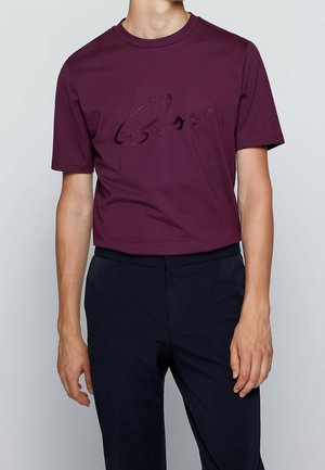 Homme portant une chemise à manches courtes couleur prune avec un logo "Boss" discret en relief, associée à un pantalon noir, sur un fond uni.