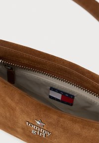 Brūns zamšs soma ar rāvējslēdzēja aizdari. Iekšpusei ir gaiša oderējums un birka ar uzrakstu "TOMMY JEANS" un sarkanā, baltā un zilā krāsā.