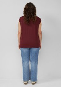 Bordeaux kleurig T-shirt met korte mouwen, losse pasvorm, gecombineerd met lichtblauwe flare jeans en beige schoeisel. Achtergrond is neutraal grijs.