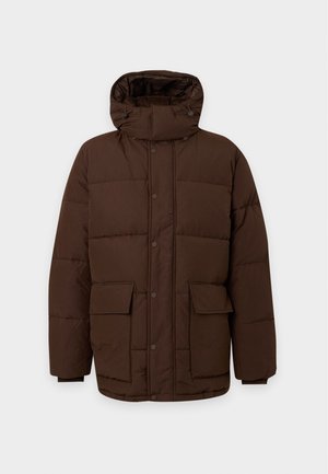 Homme portant une doudoune marron foncé, un jean bleu foncé et des baskets marron clair, debout avec une main dans la poche sur fond blanc.