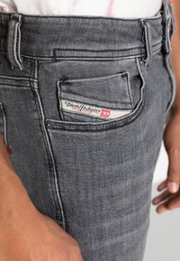 Primer plano de una persona que lleva unos vaqueros de denim Diesel Industry de color gris oscuro, con una etiqueta de marca visible en el pequeño bolsillo delantero.