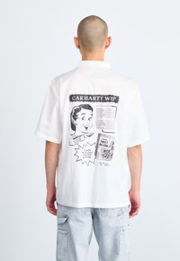 Camisa blanca de manga corta con un diseño gráfico en la parte posterior que incluye texto negro e ilustraciones. Corte relajado con cuello y dobladillo recto.