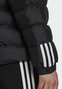 Doudoune noire à texture matelassée, poignets élastiques avec des rayures blanches, et un design élégant et chaud.