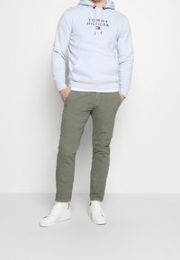 Sudadera con capucha azul claro con un logo, combinada con pantalones tapered verde oliva y zapatillas blancas. Cuenta con un bolsillo frontal y cordones.