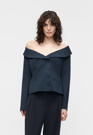 Woman wearing a dark off-shoulder buttoned blazer with long sleeves and matching dark trousers against a plain background.