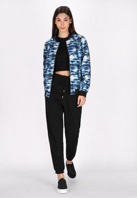 Blå og hvid mønstret bomberjakke over en sort crop top, parret med sorte sweatpants og sorte sneakers med hvide såler.