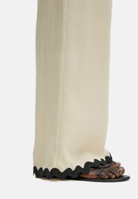 Zwarte sandalen met dunne banden, gedragen met crèmekleurige broek met een golvende zwarte rand aan de zoom, die een gladde stofstructuur toont.
