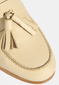 Cremefarbene Lederloafers mit Fransenquasten, runder Zehenform, sichtbarem Nähen und weicher Textur. Flache beige Sohle.
