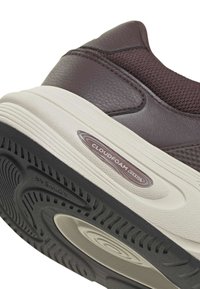 Gros plan sur le talon d'une basket marron foncé avec une semelle blanche, avec une étiquette de coussin "Cloudfoam" et un motif de semelle noire texturé.