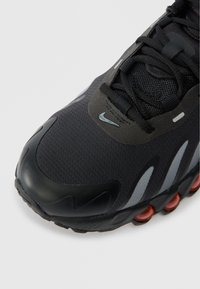 Czarny but sportowy z siateczkową cholewką, odblaskowymi paskami, czarnymi sznurówkami i widocznym czerwonym amortyzatorem w podeszwie.