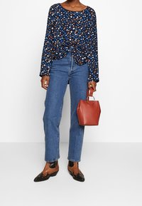 Top floreale blu navy, taglio morbido, maniche lunghe; jeans dritti azzurri chiari; scarpe marroni e nere; borsa in pelle rossa con manico in metallo.