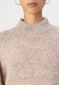 Beige high-neck sweater with a soft, textured knit, featuring a ribbed collar and relaxed fit. The fabric appears warm and fuzzy.
