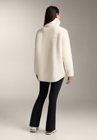 Giacca in pile color crema con colletto alto, vestibilità rilassata e tessuto texturizzato. Abbinata a pantaloni neri a zampa d'elefante e sneakers grigie.