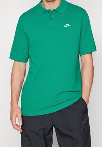 Man i en grön kortärmad polo skjorta från Nike och svarta cargobyxor från Nike, stående med ena handen i fickan mot en neutral bakgrund.