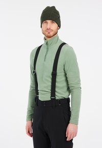 Fleece pullover in lichtgroen met een ritskraag, gecombineerd met zwarte verstelbare broekbanden en een broek. De hoed is donkergroen gebreid.