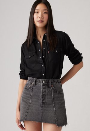 Camicia nera con bottoni e due tasche sul petto, abbinata a una gonna in denim grigia a vita alta con orlo sfrangiato e tasche frontali.